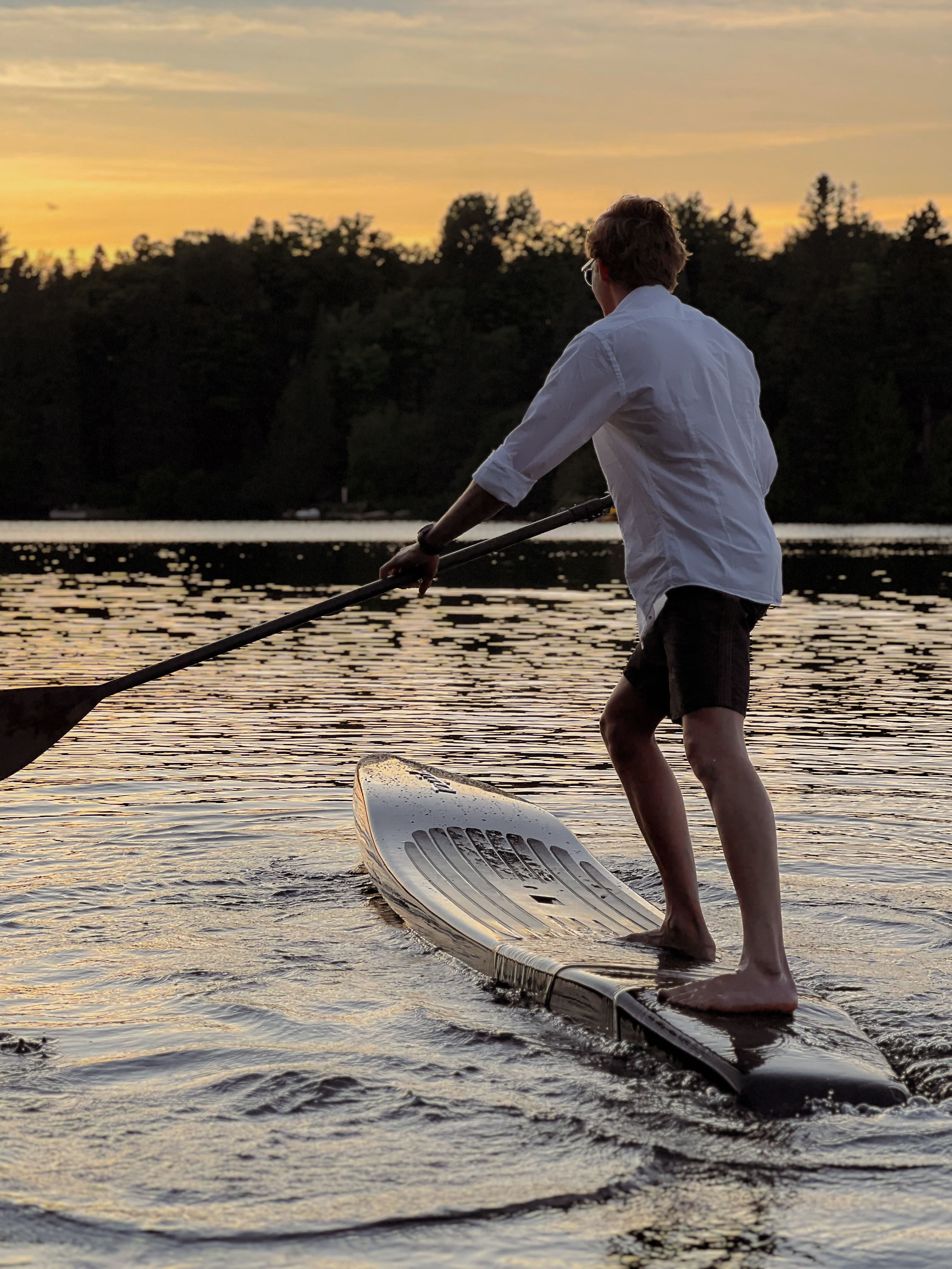 Man paddleboarding on a lake at sunset, wearing a white shirt and using a wooden paddle on a Beau Lake Rapid board