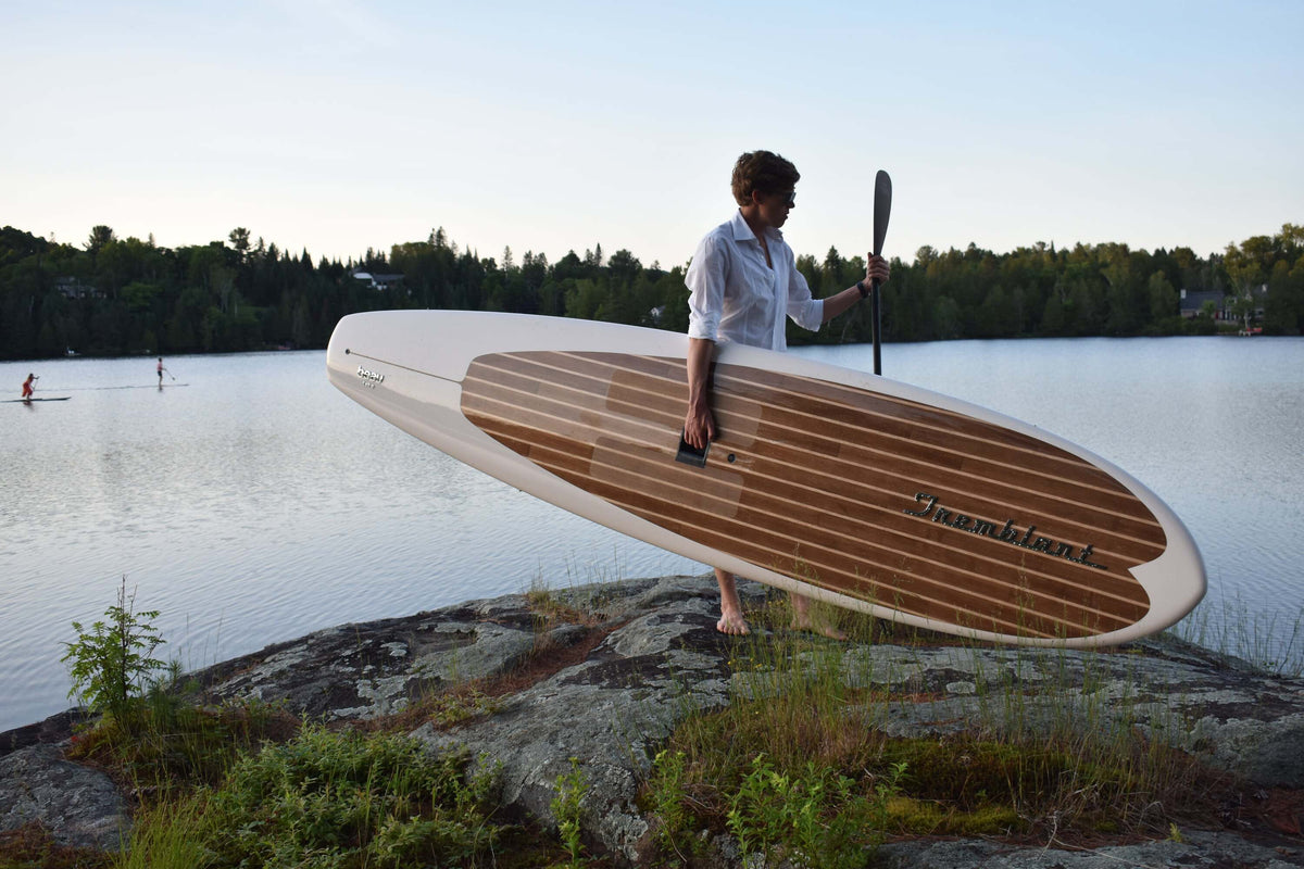 Man holding a Tremblant paddleboard by Beau Lake on a rocky lakeshore, preparing to paddleboard on calm water at sunset.